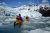 Two kayakers in orange boats with red life jackets paddle toward a towering blue glacier front in Kenai Fjords National Park.