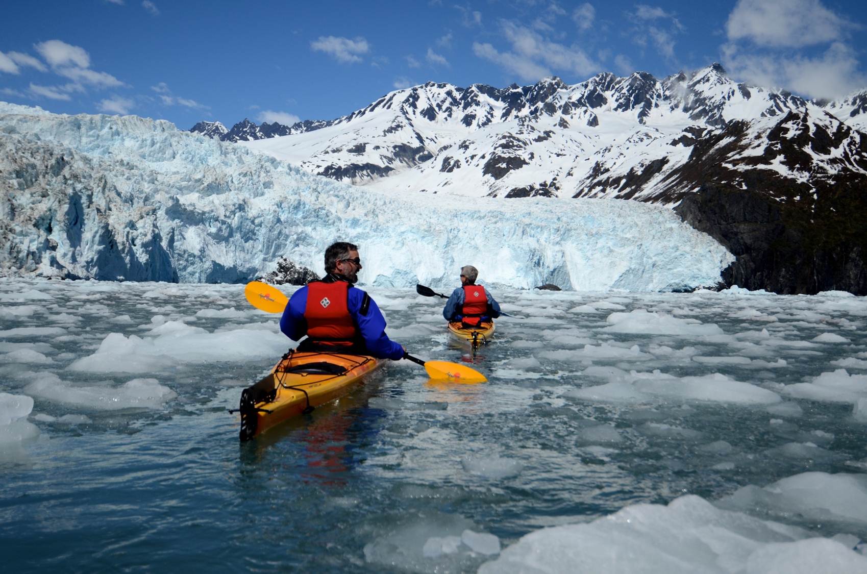 Two kayakers in orange boats with red life jackets paddle toward a towering blue glacier front in Kenai Fjords National Park.