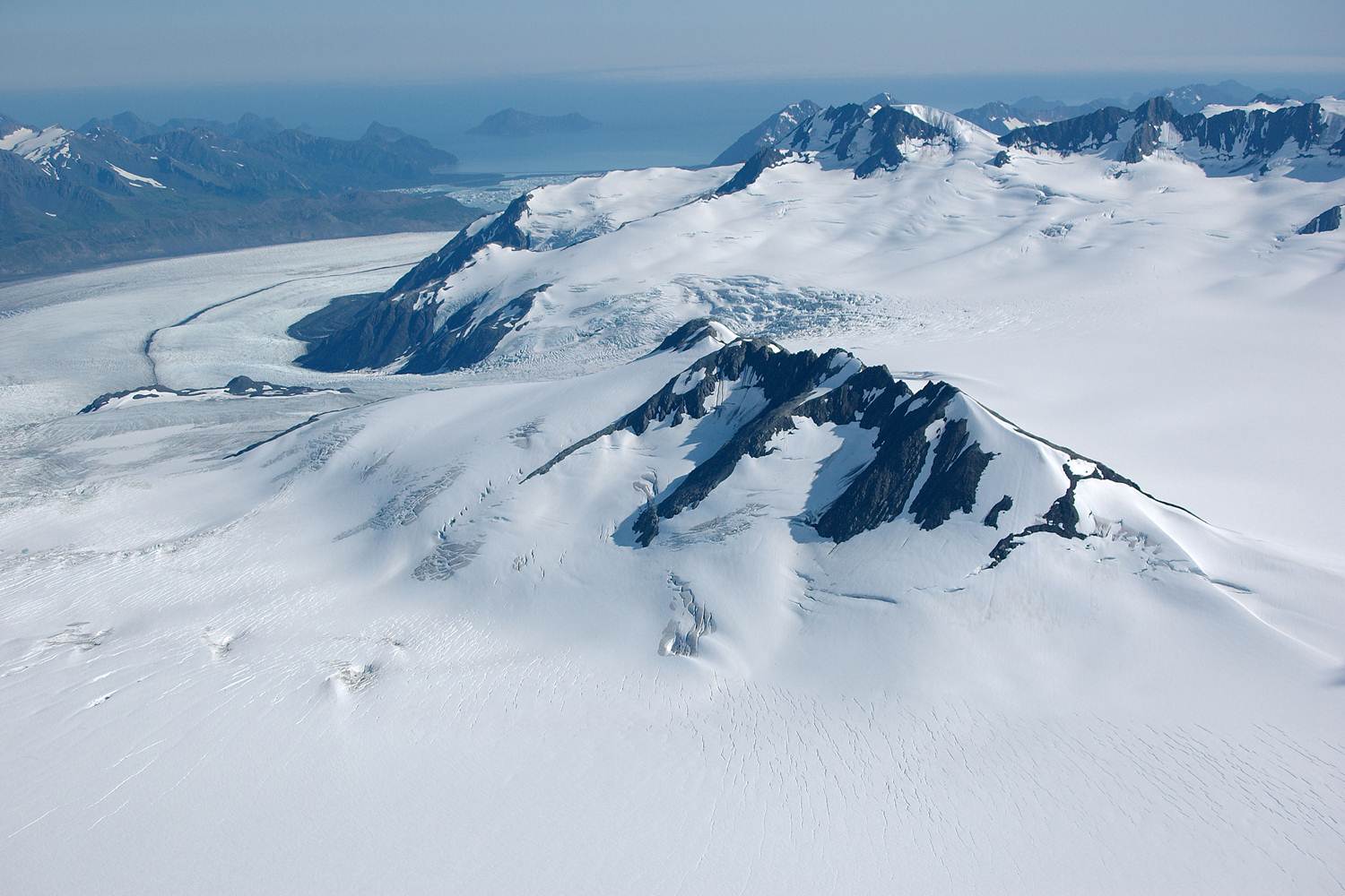 Snow-covered peaks and expansive glaciers in Kenai Fjords National Park, with a vast icefield and distant blue sea.