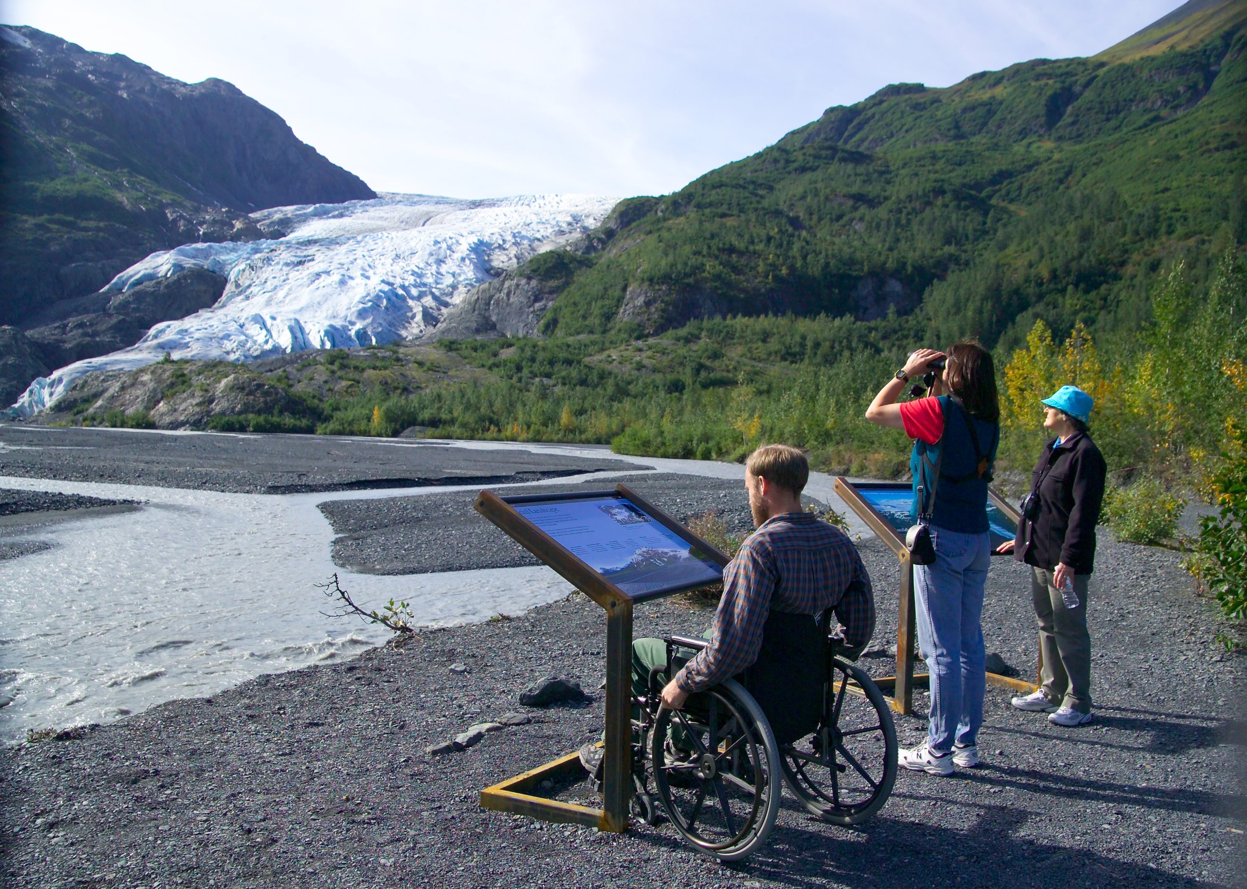 Visitors at the Exit Glacier overlook in Kenai Fjords National Park, with the glacier carving a blue tongue behind green hills.