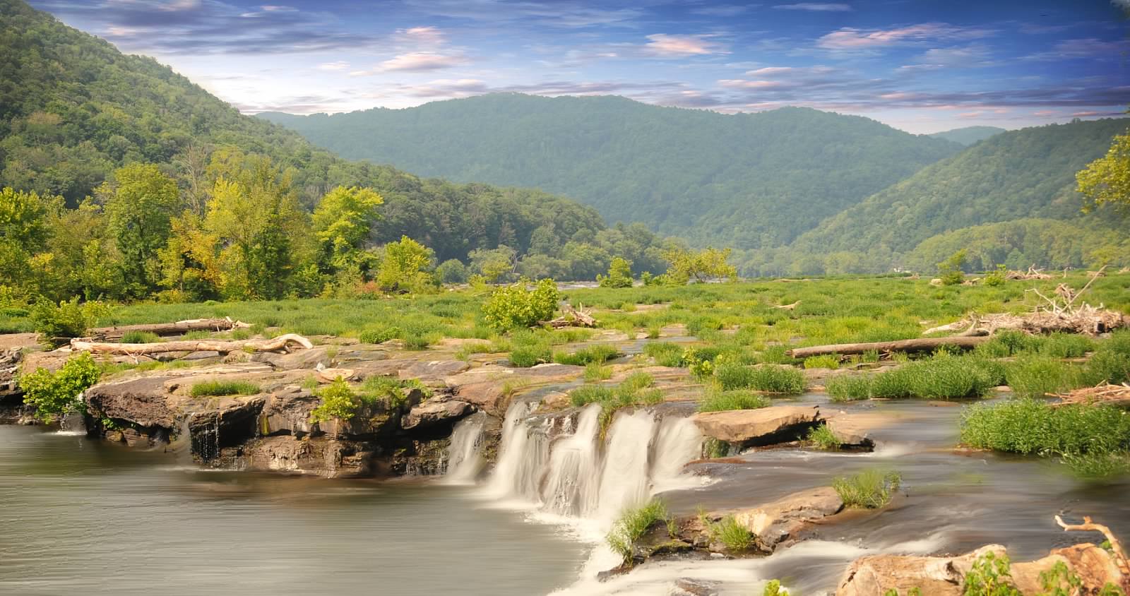 Water flows over a rocky ledge in a lush, green river valley with forested hills in the distance at New River Gorge National Park & Preserve.