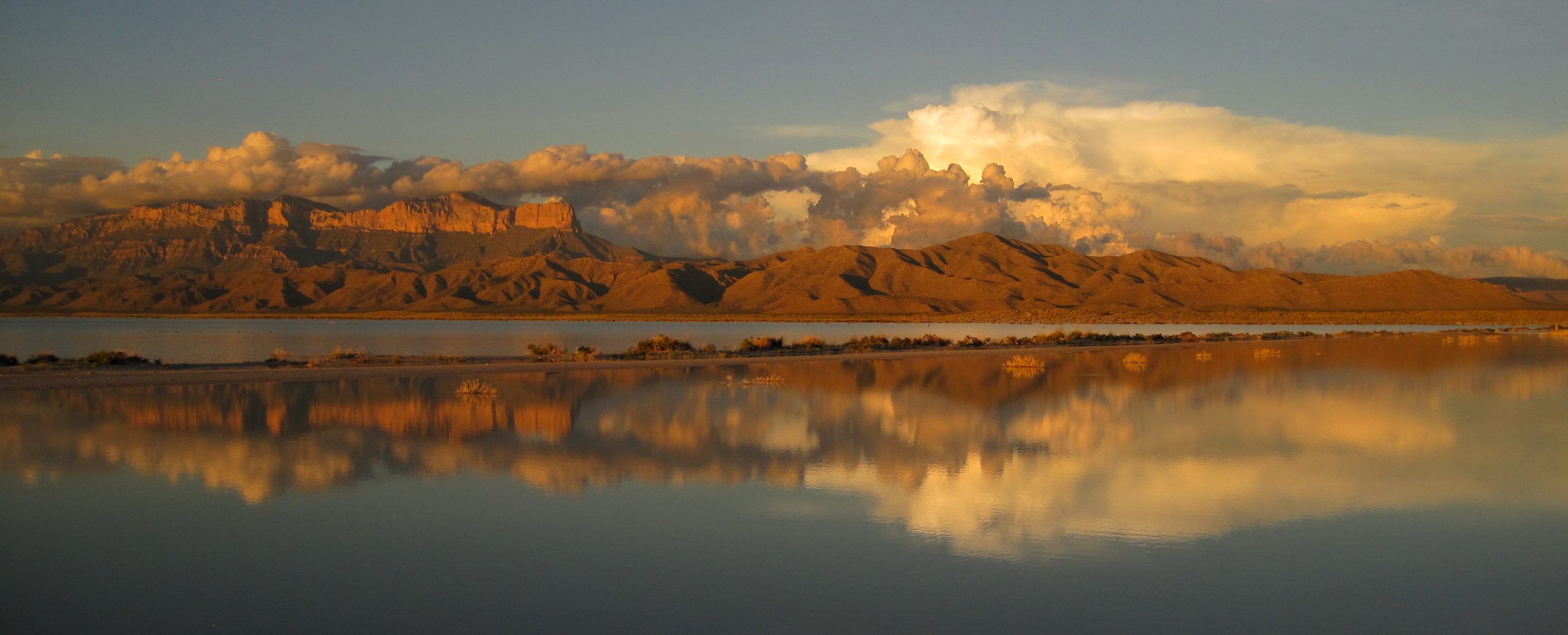 Sunset glow over the Guadalupe Mountains shows El Capitan rising above desert flats and a reflective foreground pool.