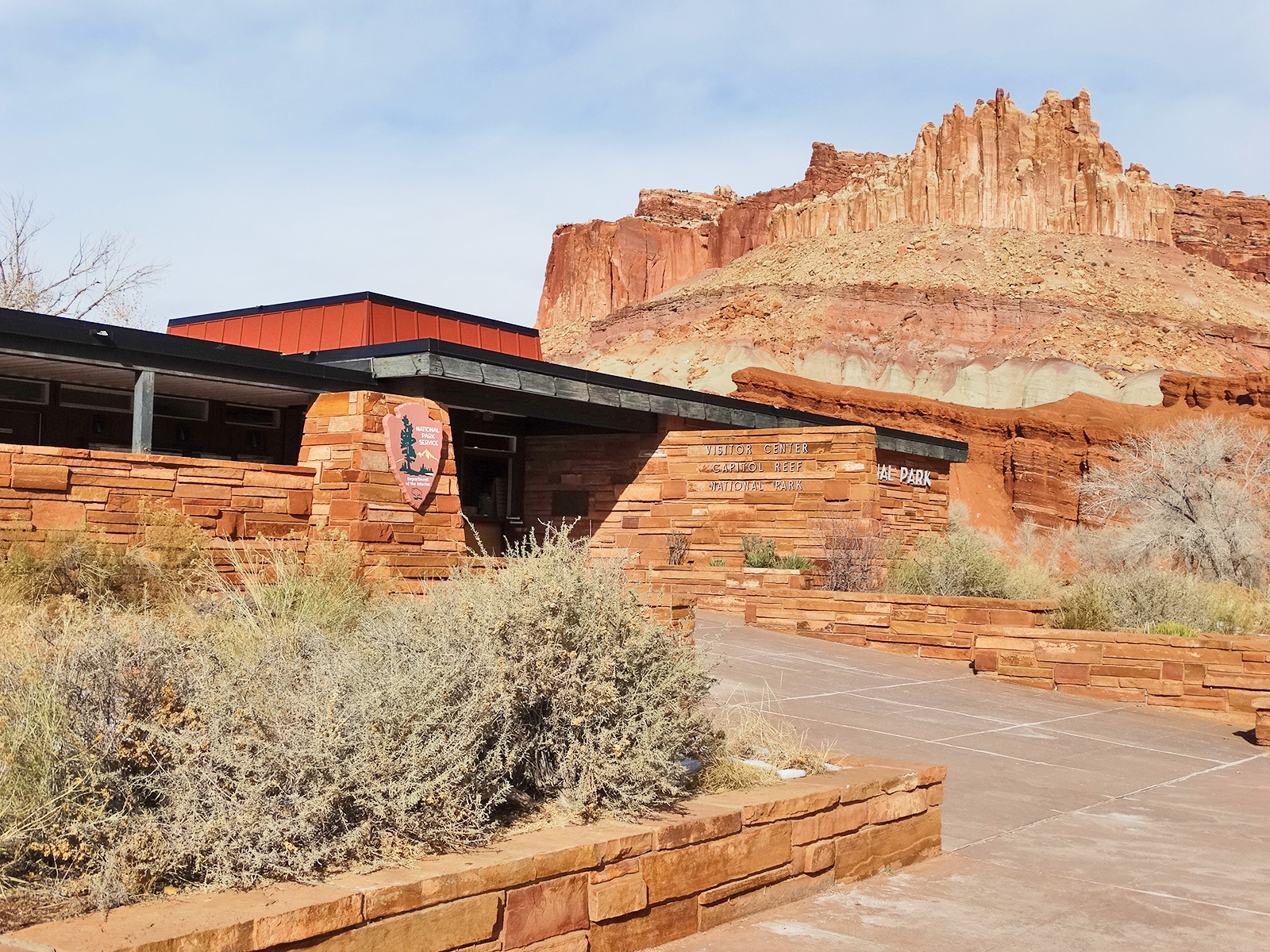 Capitol Reef National Park Visitor Center sits among red sandstone walls with a low, flat roof and desert shrubs in the foreground.
