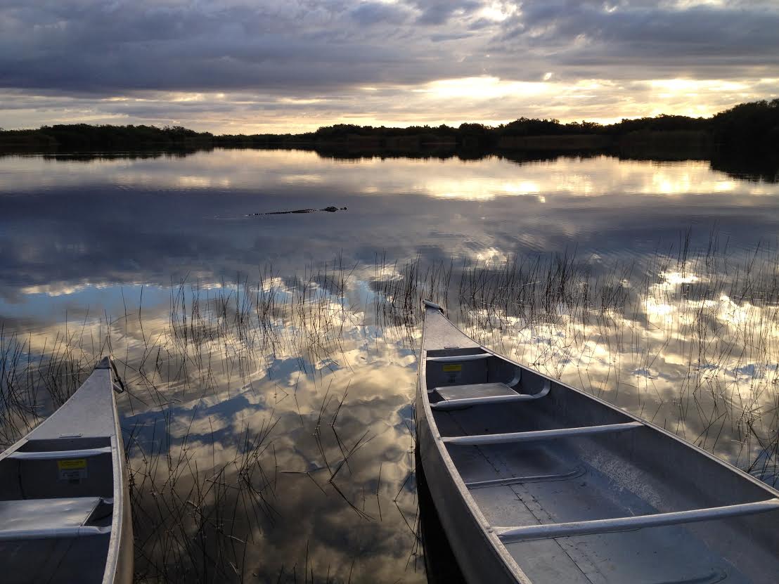 Two aluminum canoes rest in shallow marsh water at sunset in Everglades National Park, with reflections of clouds and grasses on the glassy surface.
