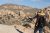 Traveler surveys a vast rocky desert panorama in Joshua Tree National Park, standing on large granite boulders with a backpack and straw hat.