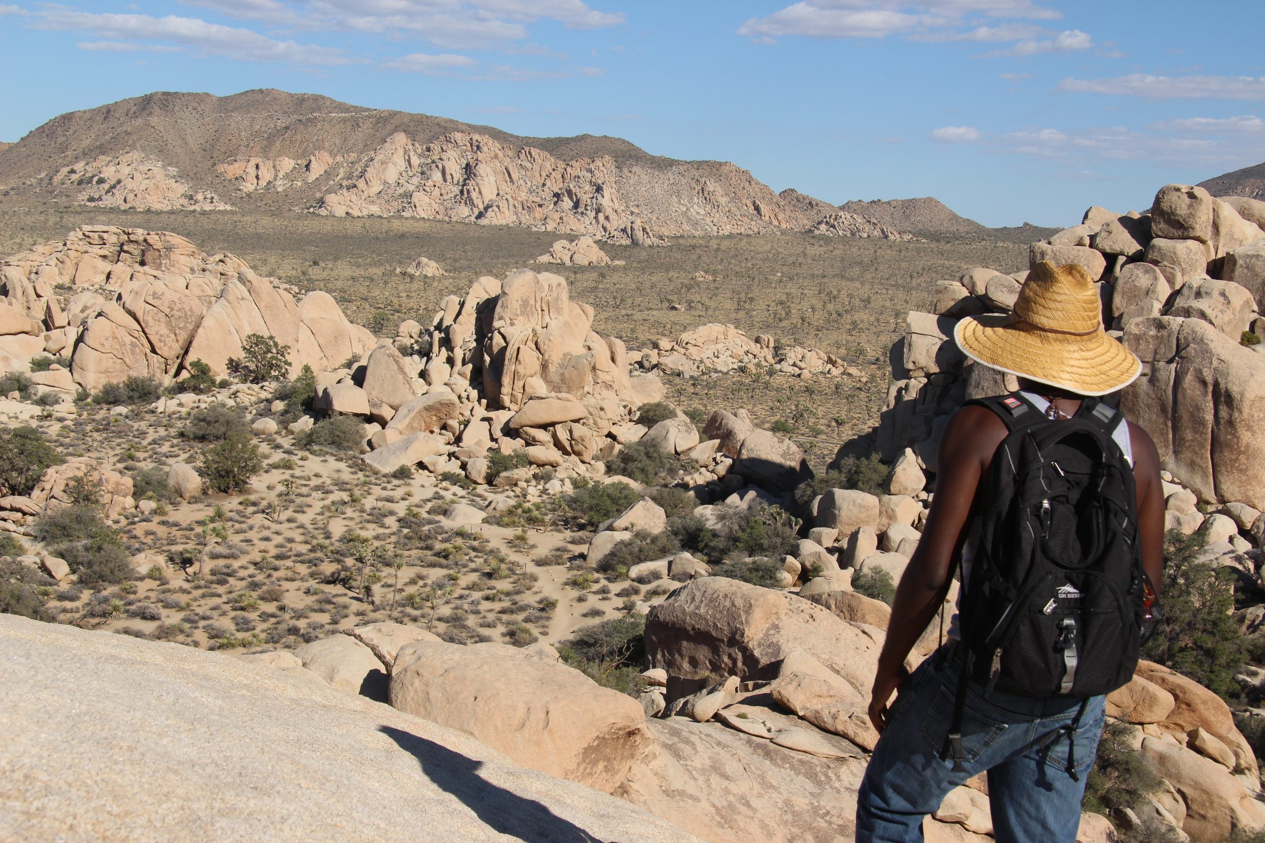 Traveler surveys a vast rocky desert panorama in Joshua Tree National Park, standing on large granite boulders with a backpack and straw hat.
