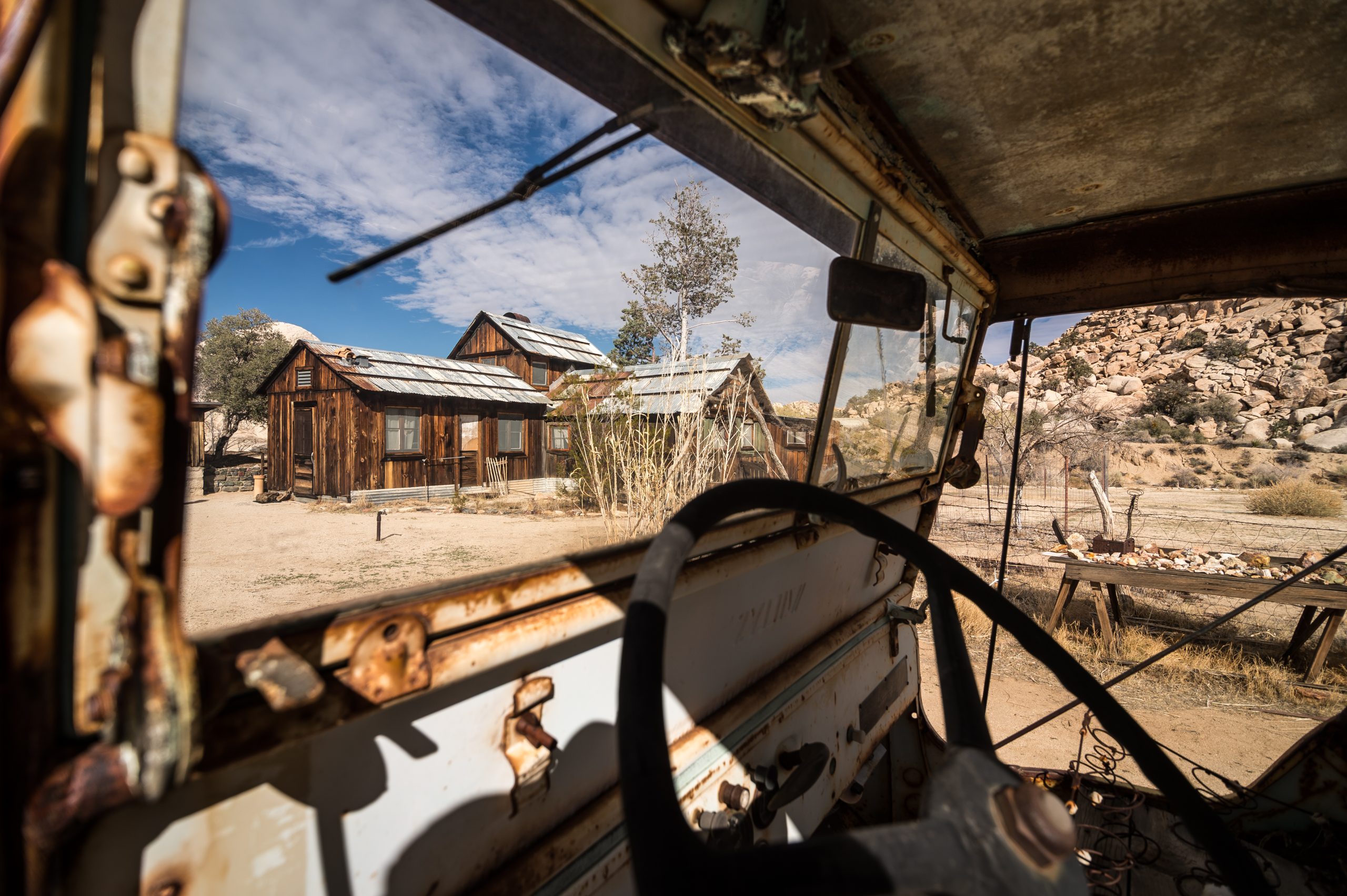 Keys Ranch in Joshua Tree National Park seen through the rusted door frame of an old vehicle, with weathered wooden cabins in the desert.