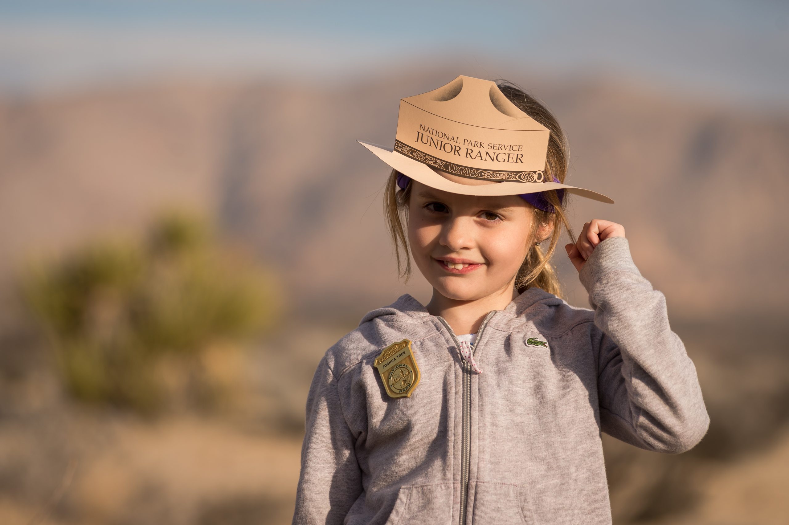 A young Junior Ranger in a tan hat stands in Joshua Tree National Park with a desert backdrop and distant rock formations.