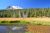 Lassen Peak rises above a green forest and a golden meadow with a calm pond in the foreground under a bright blue sky in Lassen Volcanic National Park.