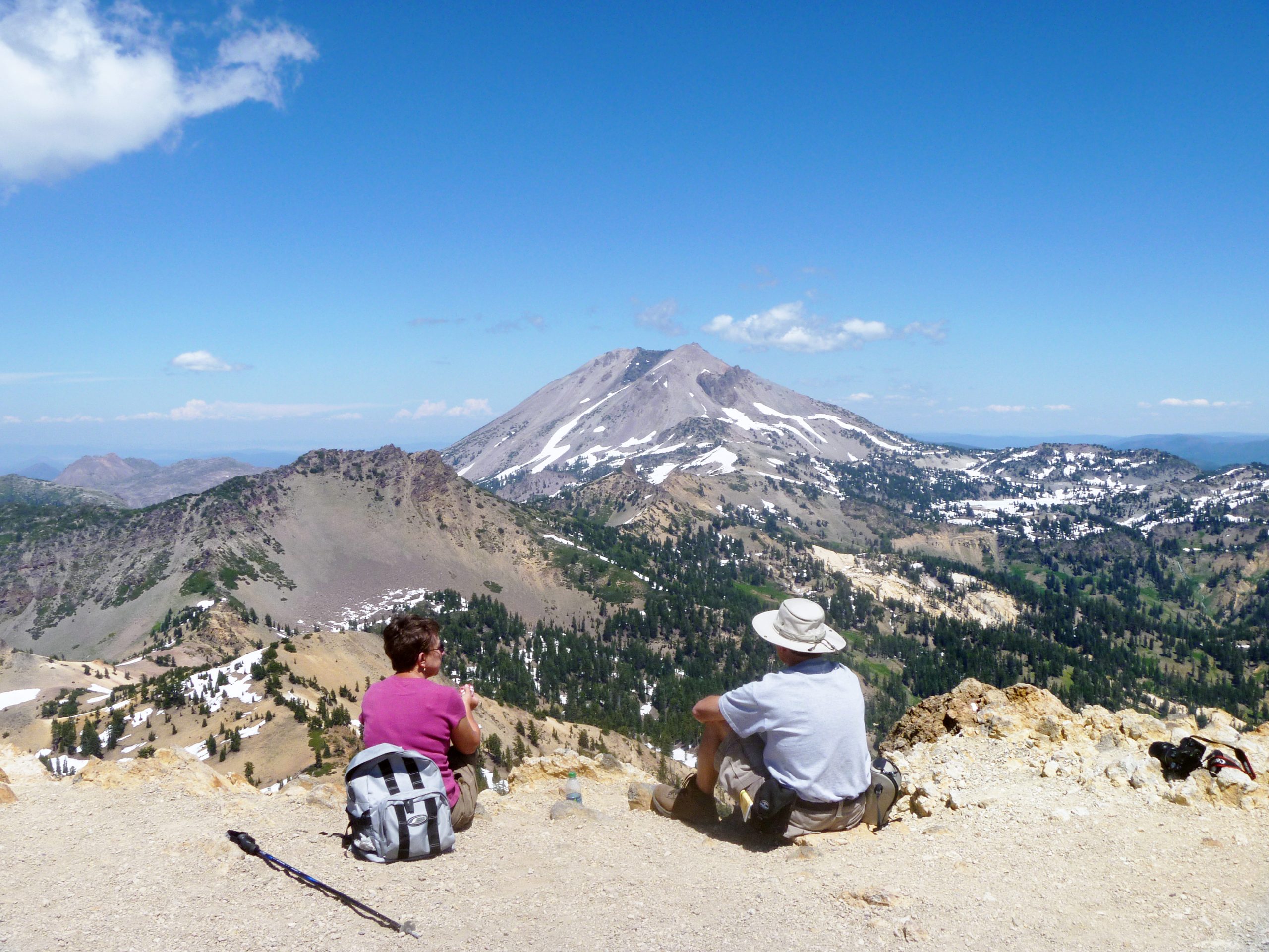 Two hikers sit on a rocky overlook in Lassen Volcanic National Park, with Lassen Peak snow-dusted and towering in the distance.