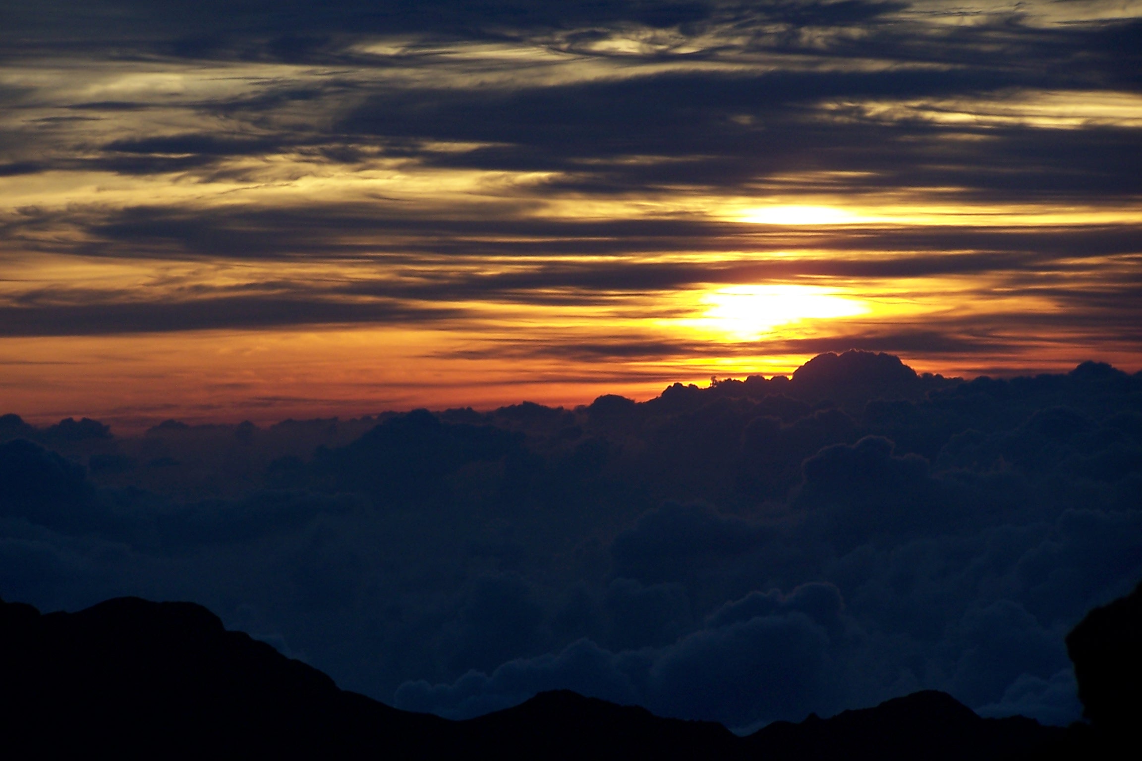 Haleakalā sunrise over a sea of clouds from the summit, with warm orange horizon and silhouetted volcanic ridges.