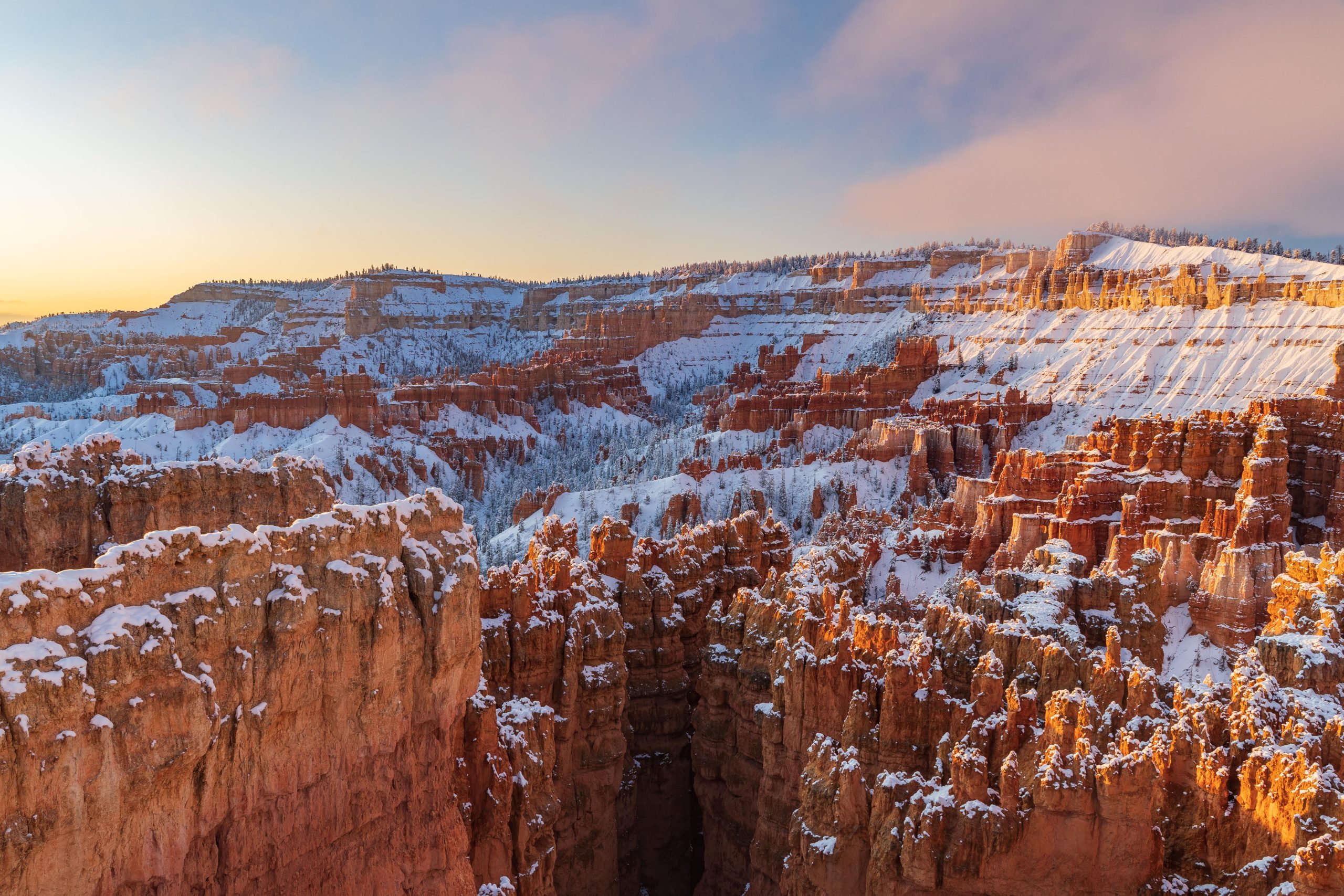 Sunlit Bryce Canyon National Park hoodoos rise from snowy cliffs along the rim at sunrise in winter light.