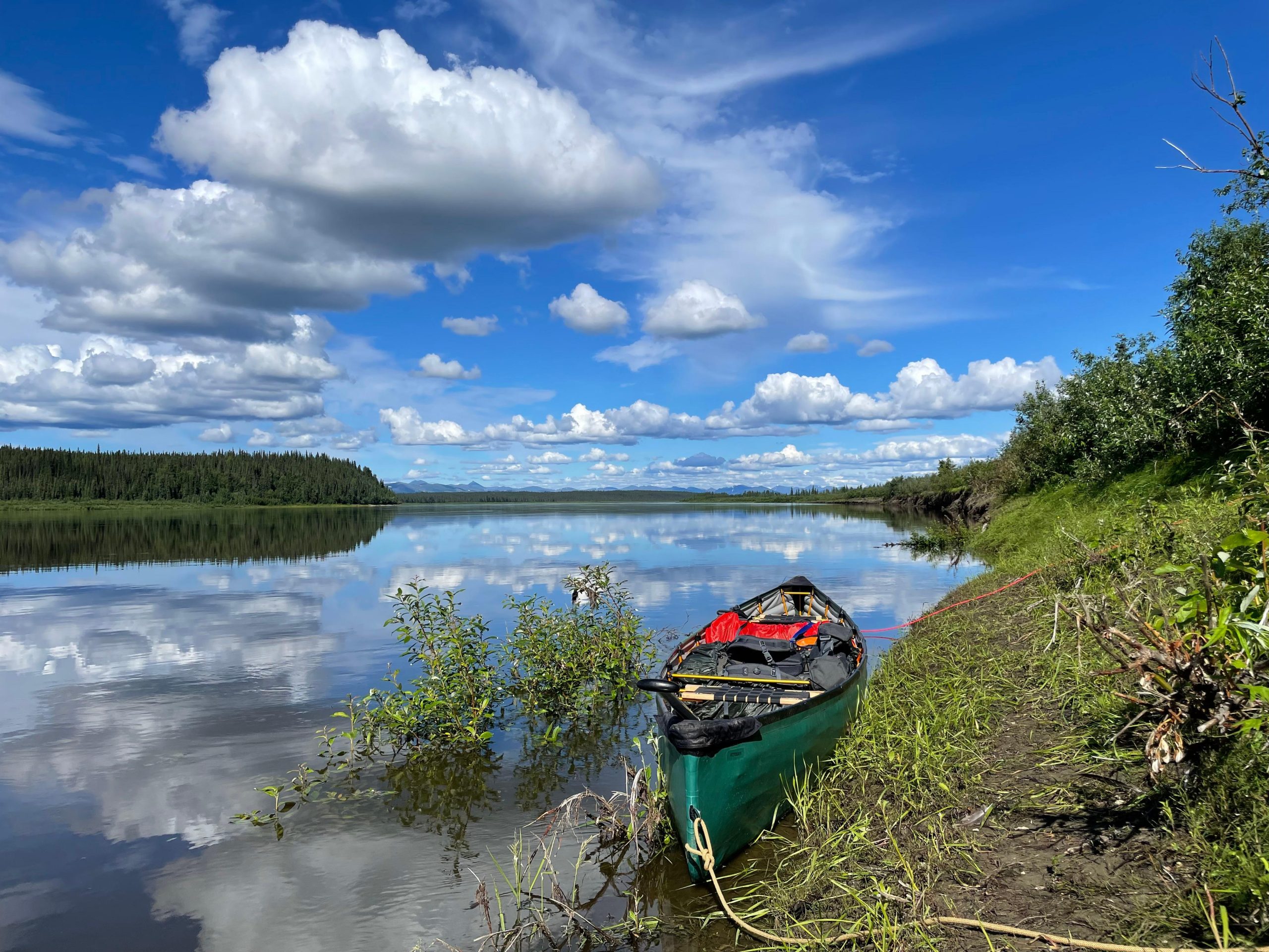 Green canoe lies tethered on the grassy Kobuk Riverbank at Kobuk Valley National Park, Alaska, with a reflective blue sky and alders along the shore.