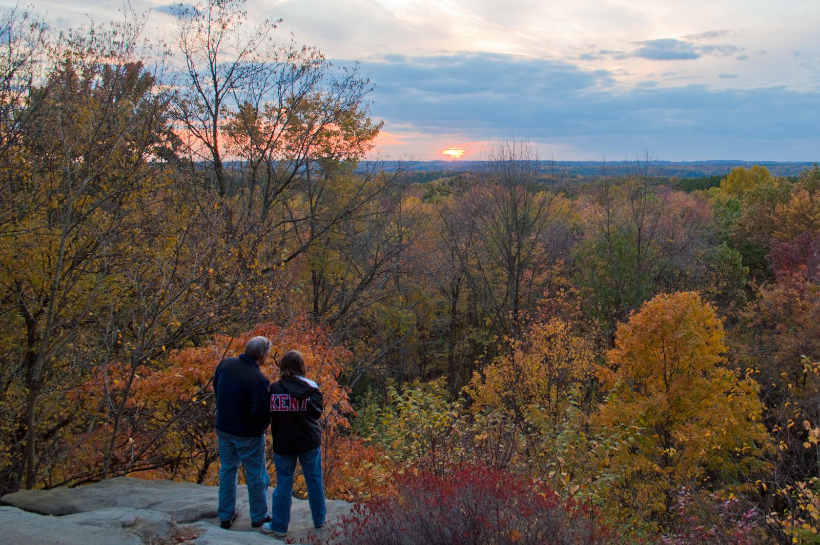 Two people stand on a rocky overlook in fall colors, watching a sunset over the forested valley in Cuyahoga Valley National Park.