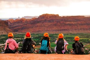 Five hikers with orange helmets sit on a rim overlooking red cliffs and a green valley in Arches National Park.