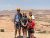 Family wearing orange helmets and harnesses stands at a sandstone overlook in Arches National Park, with red rock mesas.