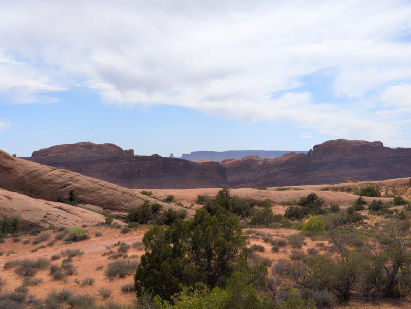 Red sandstone mesas and desert shrubs spread across a wide canyon under a partly cloudy blue sky in Arches National Park.
