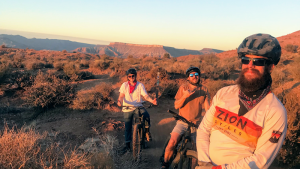 Three cyclists ride a desert trail near Zion National Park at sunset, with rugged mesas on the horizon.