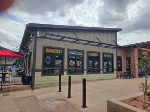 Exterior of a small modern restaurant with neon signs and outdoor seating along a sidewalk in Zion National Park.