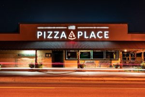 Pizza Place storefront on a Bryce Canyon National Park street at night with bright neon sign and starry sky above.