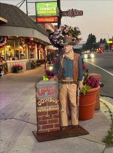 Bryce Canyon National Park street scene featuring a life-size cowboy statue beside a brick sign for a smokehouse cafe, with flowers and a storefront in the background.