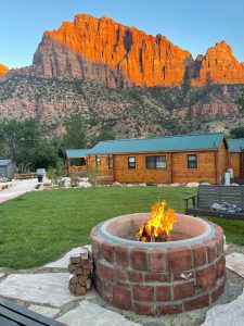 Zion Canyon Campground cabins with a stone fire pit against the orange cliffs of Zion National Park.