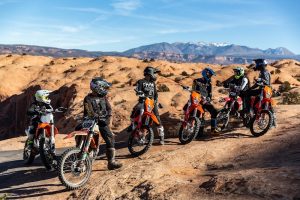 Group of riders on orange dirt bikes pause on a dusty Arches National Park trail amid red rock formations.