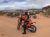 Motorcycle rider in an orange dirt bike and protective gear rests on a sandy track in Arches National Park, with desert scrub and distant red rock formations under a partly cloudy sky.