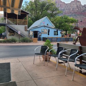 Blue cafe building with outdoor seating along a Zion National Park street, set against red rock cliffs and trees in the background.