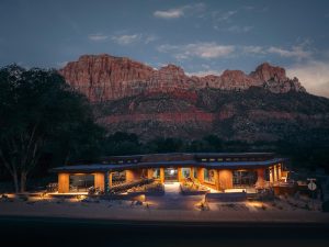 Zion Canyon Visitor Center glows warmly at dusk with towering red cliffs of Zion National Park in the background.