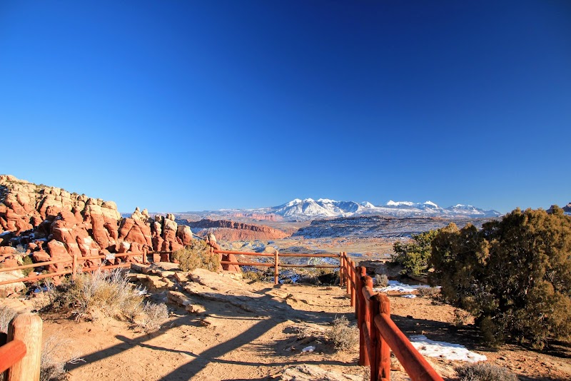 Sunlit red rock formations and a wooden railing overlook Fiery Furnace in Arches National Park, with distant snow-capped mountains.