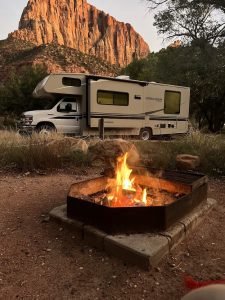 South Campground at Zion National Park features a parked motorhome and a lit campfire under towering red cliffs.