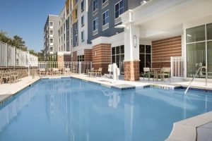 Pool deck at a modern hotel near Congaree National Park, showing a bright outdoor pool and adjacent patio area with multi-story lodging in the background.
