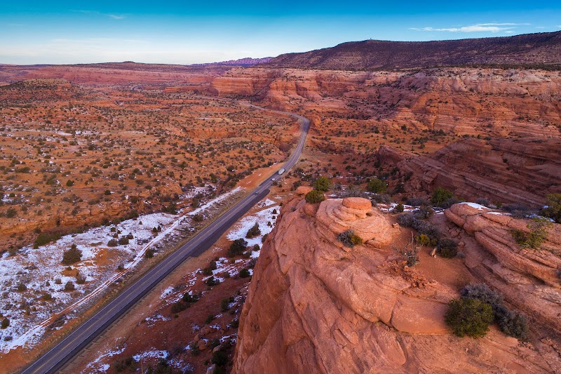 Aerial view of a winding road through red sandstone at Arches National Park, with sparse desert vegetation and snow patches.