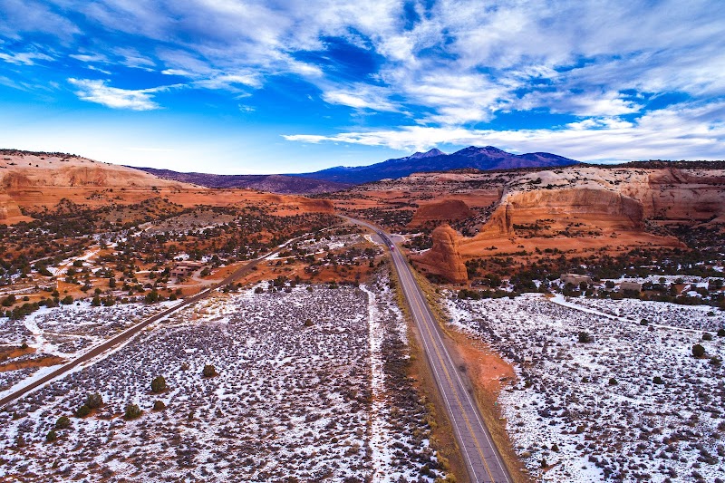Aerial view of a highway through a snow-dusted desert with red rock arches and distant mesas in Arches National Park.