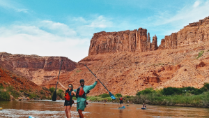 Two people in life jackets paddle on a muddy river at Arches National Park, with red canyon walls and blue sky behind.