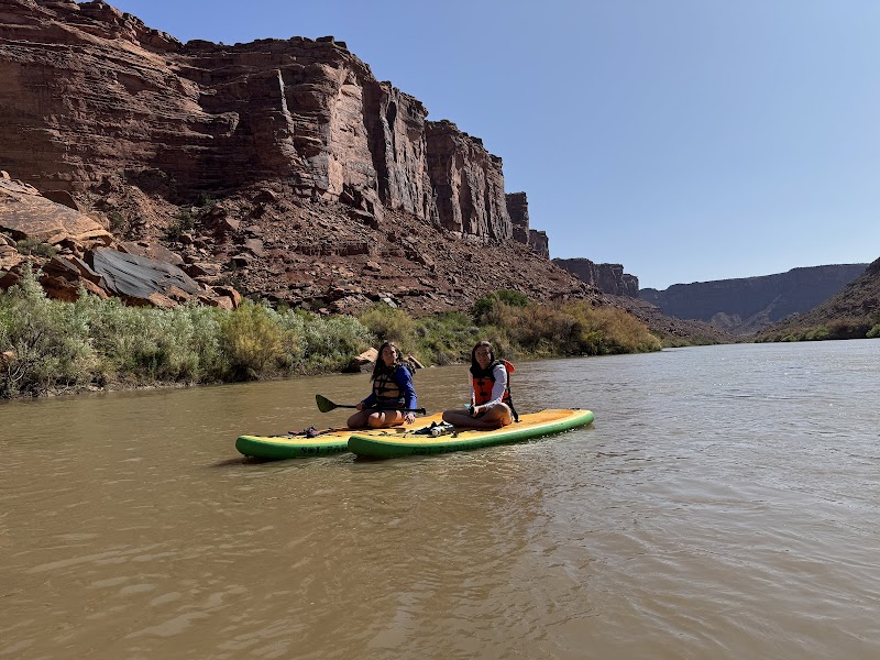 Two people in life vests paddle a bright green and yellow board down a muddy river beside towering red cliffs in Arches National Park.