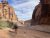 Hiker in a hat and backpack walks a sandy canyon lane between towering red sandstone cliffs at Arches National Park.