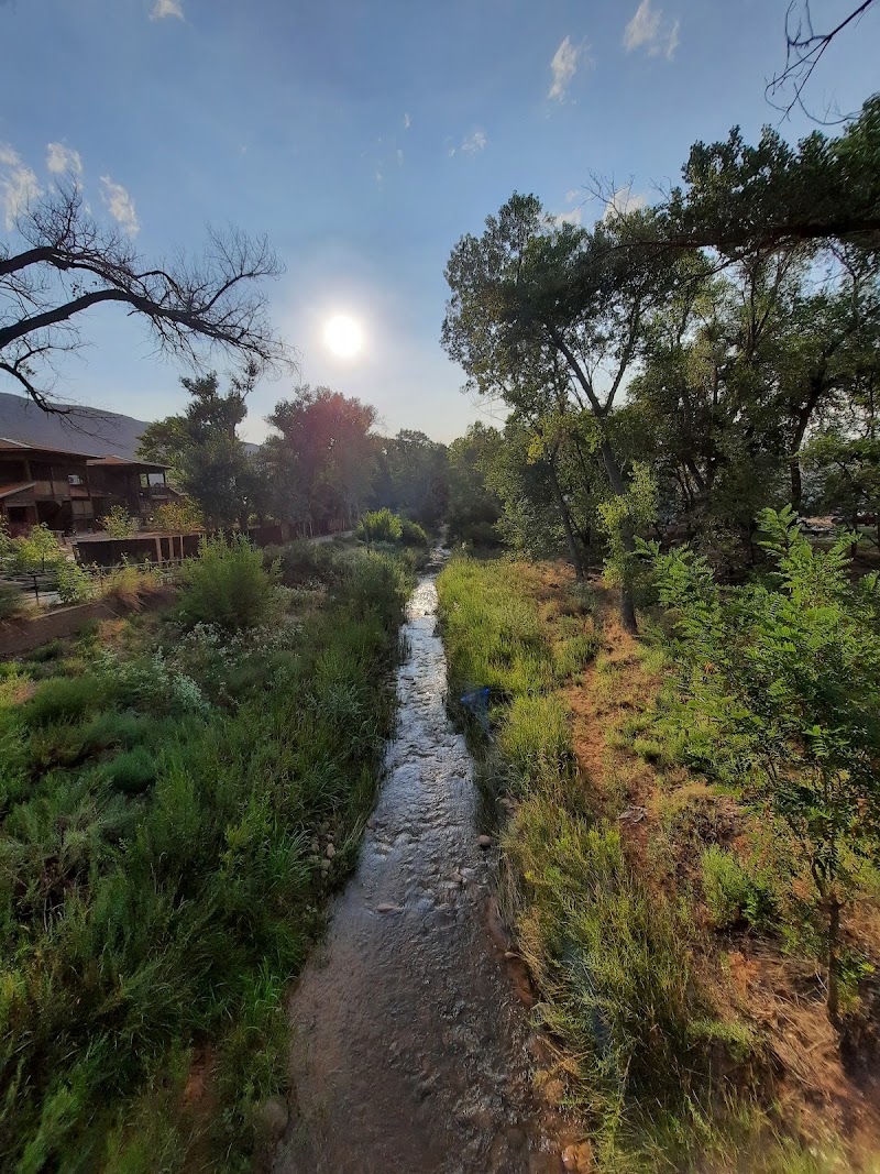 Sunlit, narrow stream winds through grassy banks and trees in Rotary Park at Arches National Park, with a wooden building on the left.