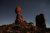 Tall balanced rock formations in Arches National Park rise over a desert floor with sparse shrubs beneath a starry night sky.