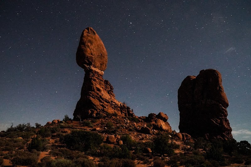 Tall balanced rock formations in Arches National Park rise over a desert floor with sparse shrubs beneath a starry night sky.