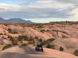 Blue off-road jeep on pink sandstone slickrock in Arches National Park, with winding trails and distant mountains.
