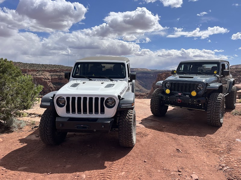 Two rugged Jeeps parked on a red dirt overlook at Arches National Park, with blue sky and canyon in the background.