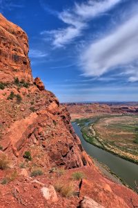 Red sandstone cliff towers on the left as a winding river cuts through the valley in Arches National Park, under a bright blue sky.