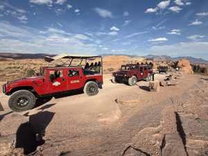 Three red open-top Jeeps drive a rocky desert trail in Arches National Park beneath a bright blue sky.