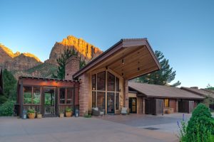 Lodge exterior with a tall stone chimney and glass entry, set against Zion Canyon cliffs at Zion National Park.