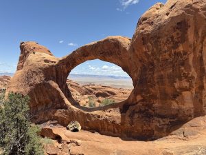 Red sandstone arch in Arches National Park frames a desert valley, blue sky, and sparse green shrubs.