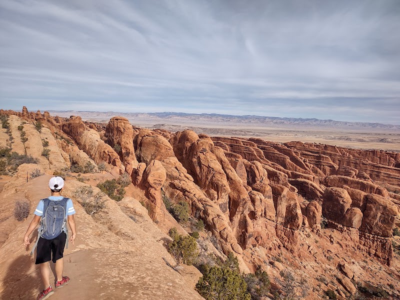 Hiker with backpack and cap walks along a dusty trail beside towering red rock formations in Arches National Park.