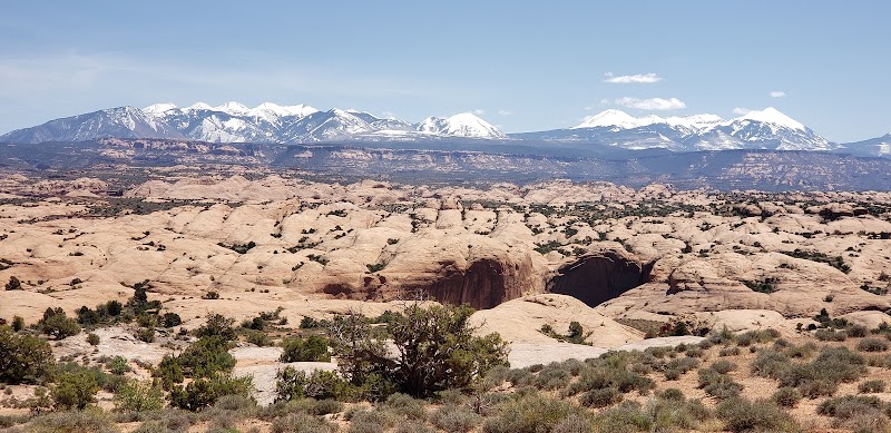Sand flats with arched rock formations and scattered shrubs stretch toward snow‑capped mountains in Arches National Park.