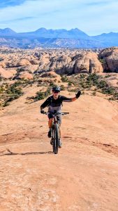 Cyclist on a fat-tire bike climbs red slickrock in Arches National Park, with orange formations and distant blue mountains.